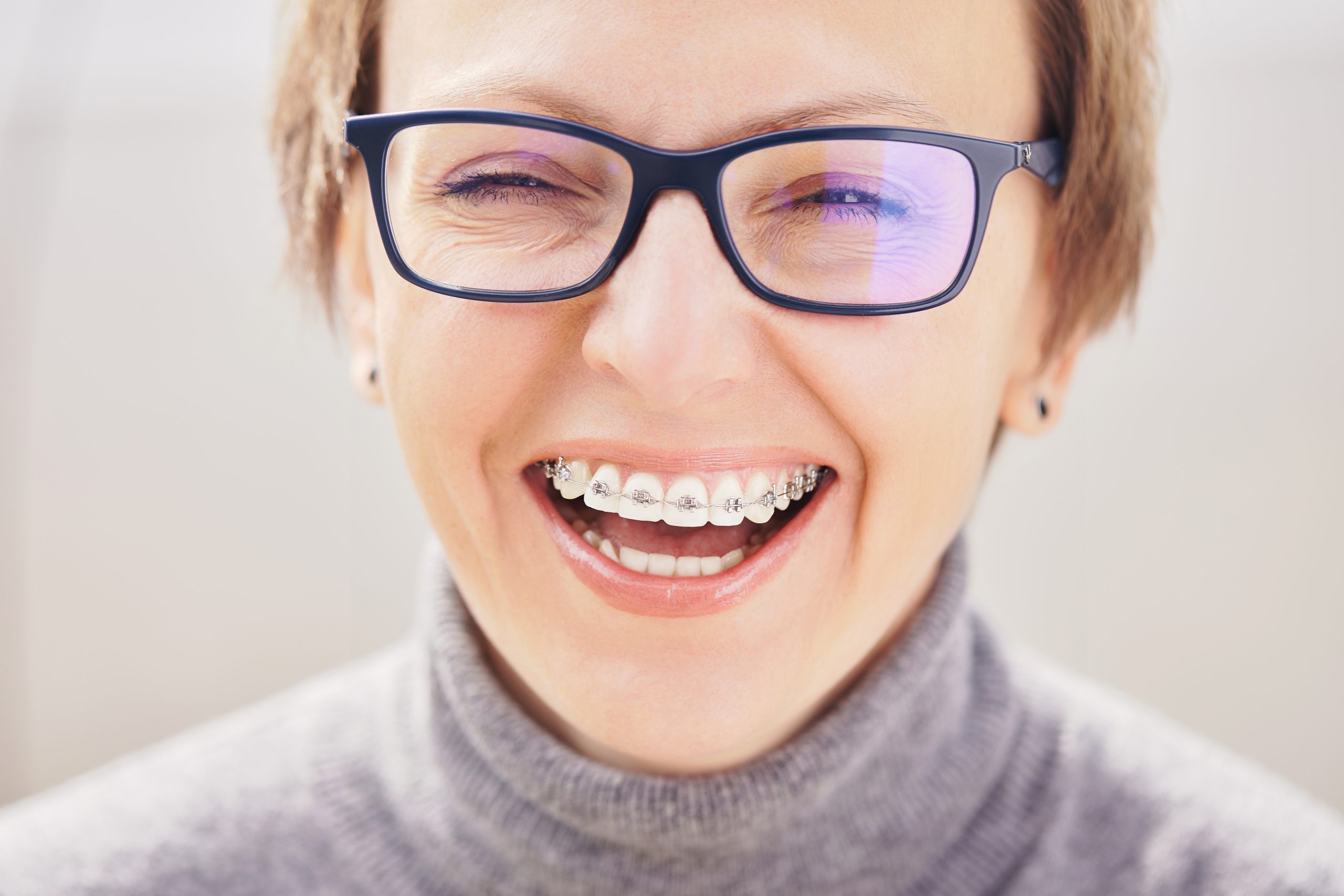 Stylish and beautiful young blonde girl in glasses and a gray sweater in braces, smiling on a white background, orthodontist dentist concept