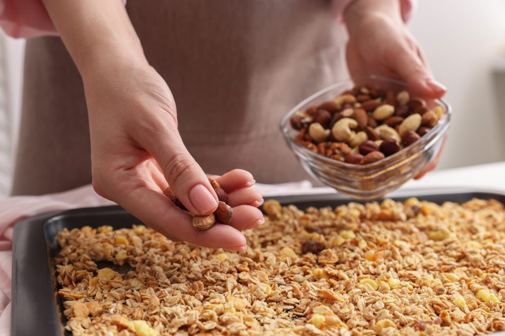 Making granola. Woman adding nuts onto baking tray with mixture of oat flakes and other ingredients at table in kitchen, closeup