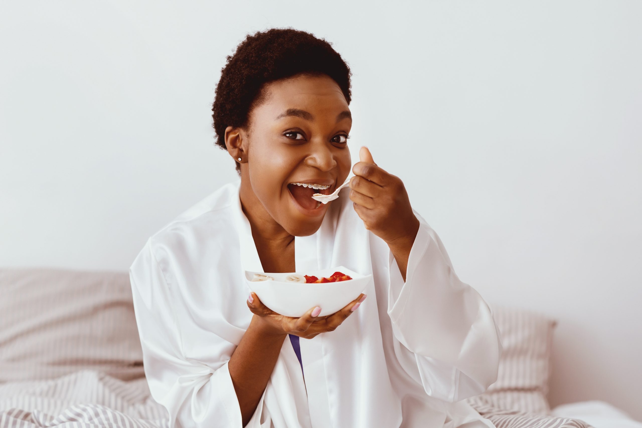Healthy eating, good morning and body care at home. Cheerful millennial african american lady in white robe with braces, eating porridge with fruit, on bed in interior of bedroom, empty space