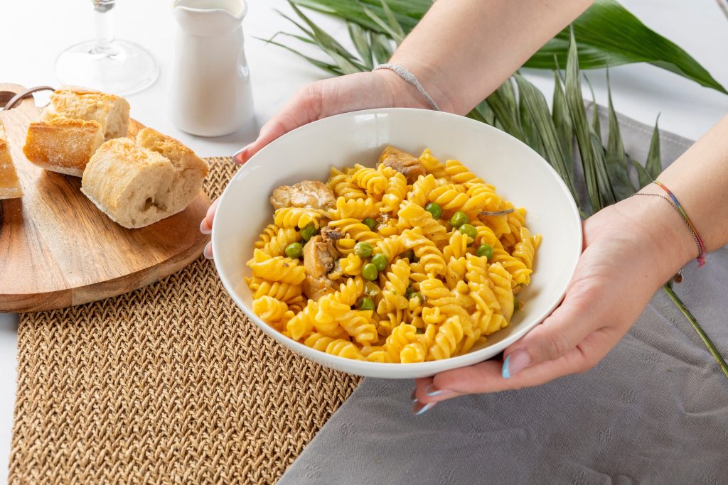 Hands holding a bowl of fusilli pasta with chicken, green peas, and mushrooms
