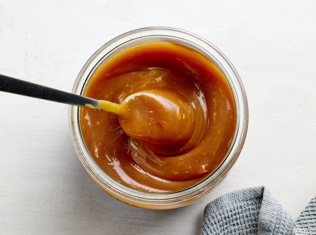 Glass jar of freshly cooked homemade caramel sauce on white kitchen table, top view