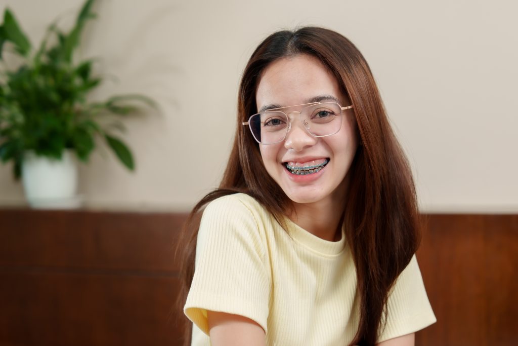 Gleeful Asian young adult with clear eyeglasses, casual yellow shirt, blue braces displayed, indoor setting. Cheerful person with spectacles, brunette hair, joyful expression
