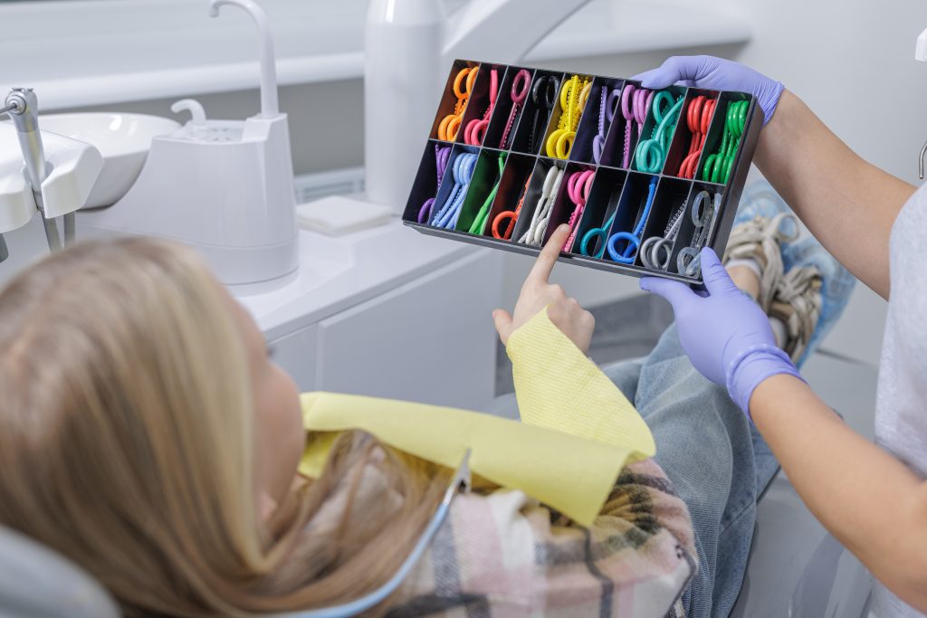 Pediatric patient pointing at colorful orthodontic ligatures in a clinic as a gloved assistant presents choices for braces treatment