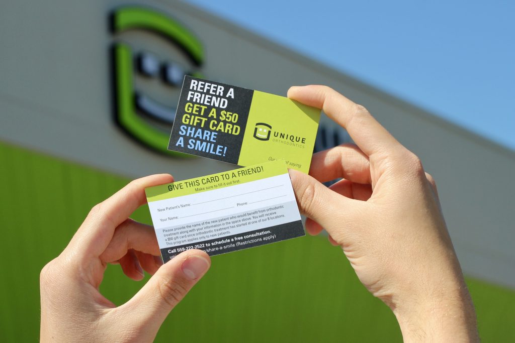 Hands holding the front and back of Unique Orthodontics’ “Share a Smile” referral cards outside the office, with the green and black Unique Orthodontics logo blurred in the background. The referral card promotes a $50 gift card for referring a friend and includes spaces for patient information on the back.