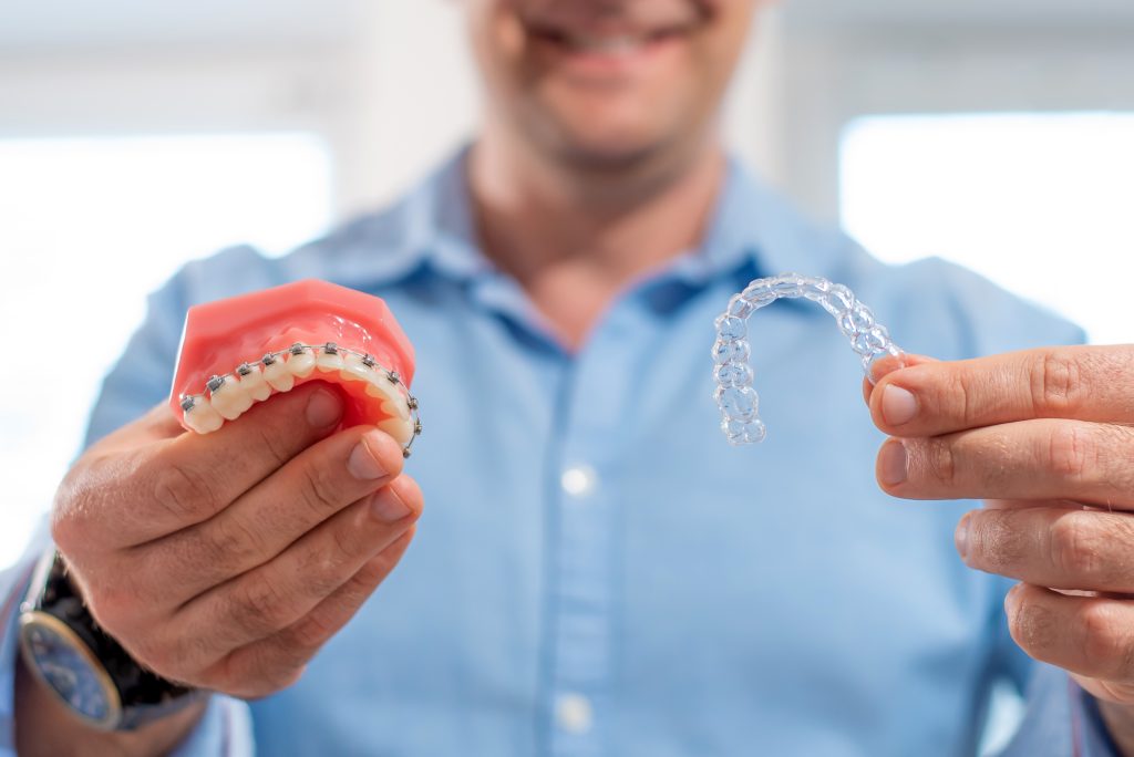 Smiling orthodontist doctor holding aligners and braces in hand shows the difference between them