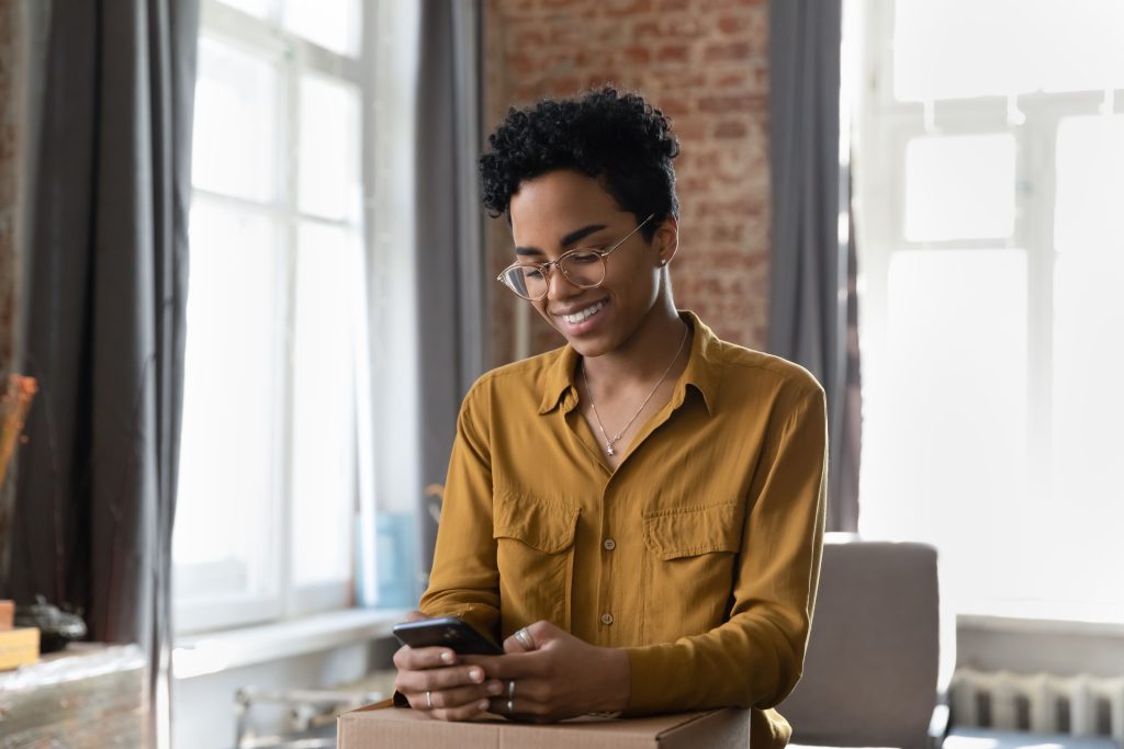Smiling attractive young African American woman in eyeglasses leaving positive review in mobile online shopping application, feeling satisfied with received internet order, recommending store.
