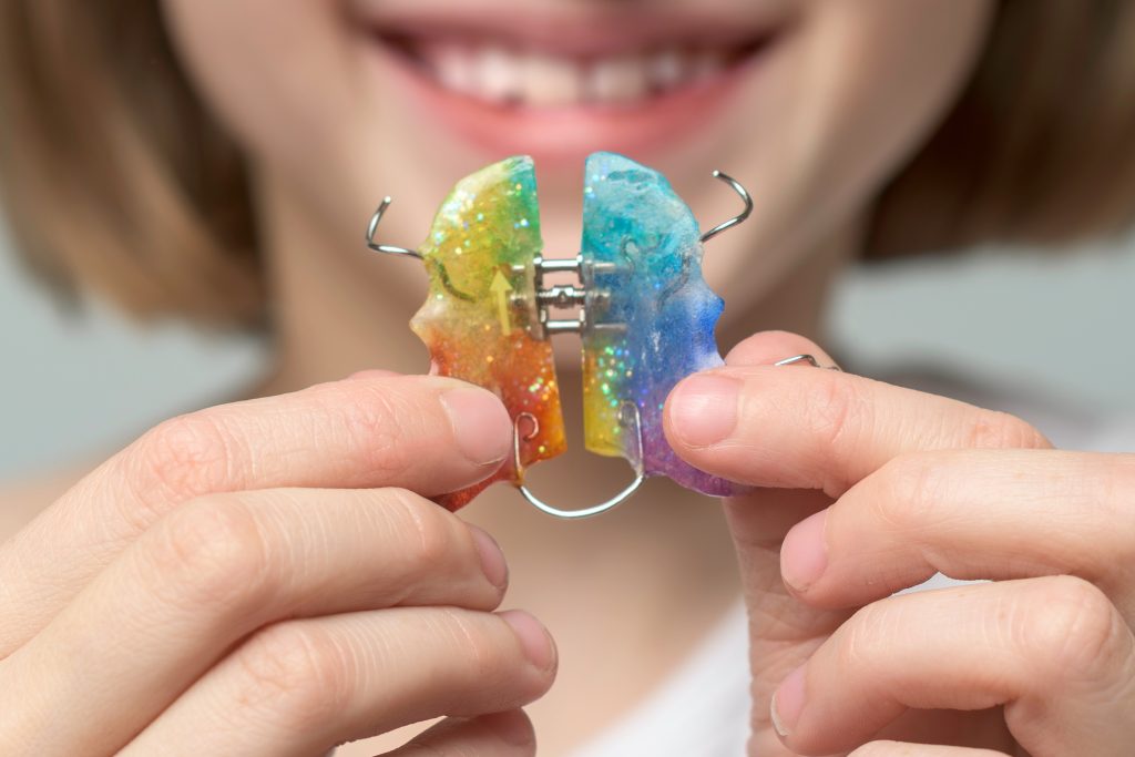 Smiling child shows colorful orthodontic an expander in hands during dental visit, bright palette adds playful mood to teeth treatment.