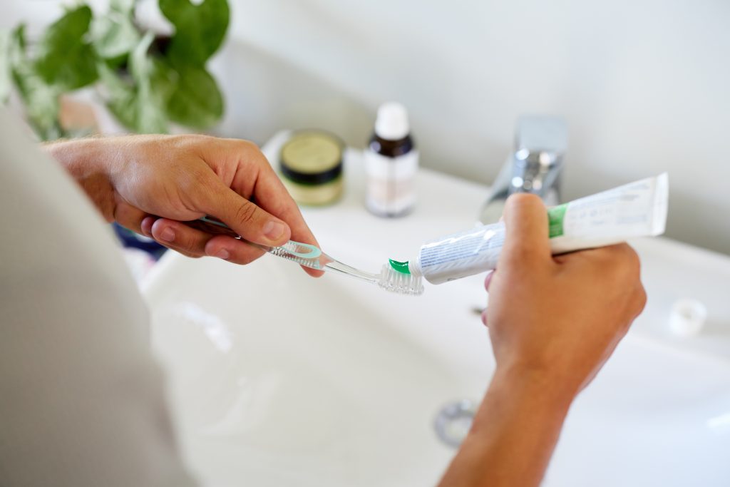 Man squeezing out toothpaste onto toothbrush before brushing. Hands gripping toothpaste tube and brush while preparing for morning routine. Man preparing for dental care routine.
