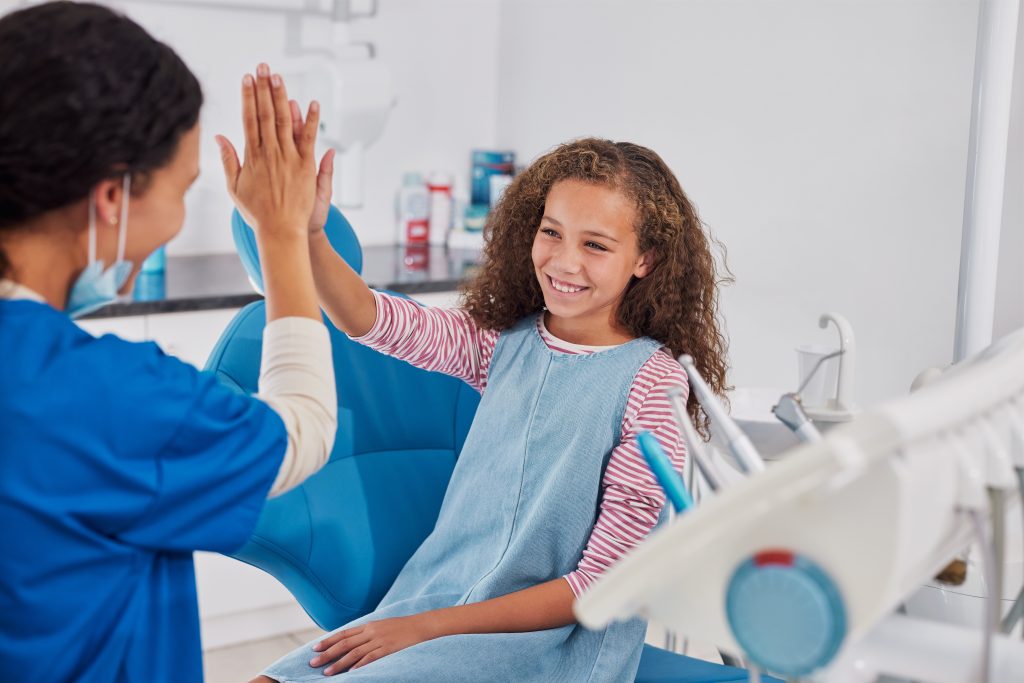 Smiling girl giving high five to dentist after dental checkup or treatment. Happy female child sit on the dental chair and cheerfully gives a high five to the nurse. Female doctor encouraging multiethnic little girl patient and giving her a high five